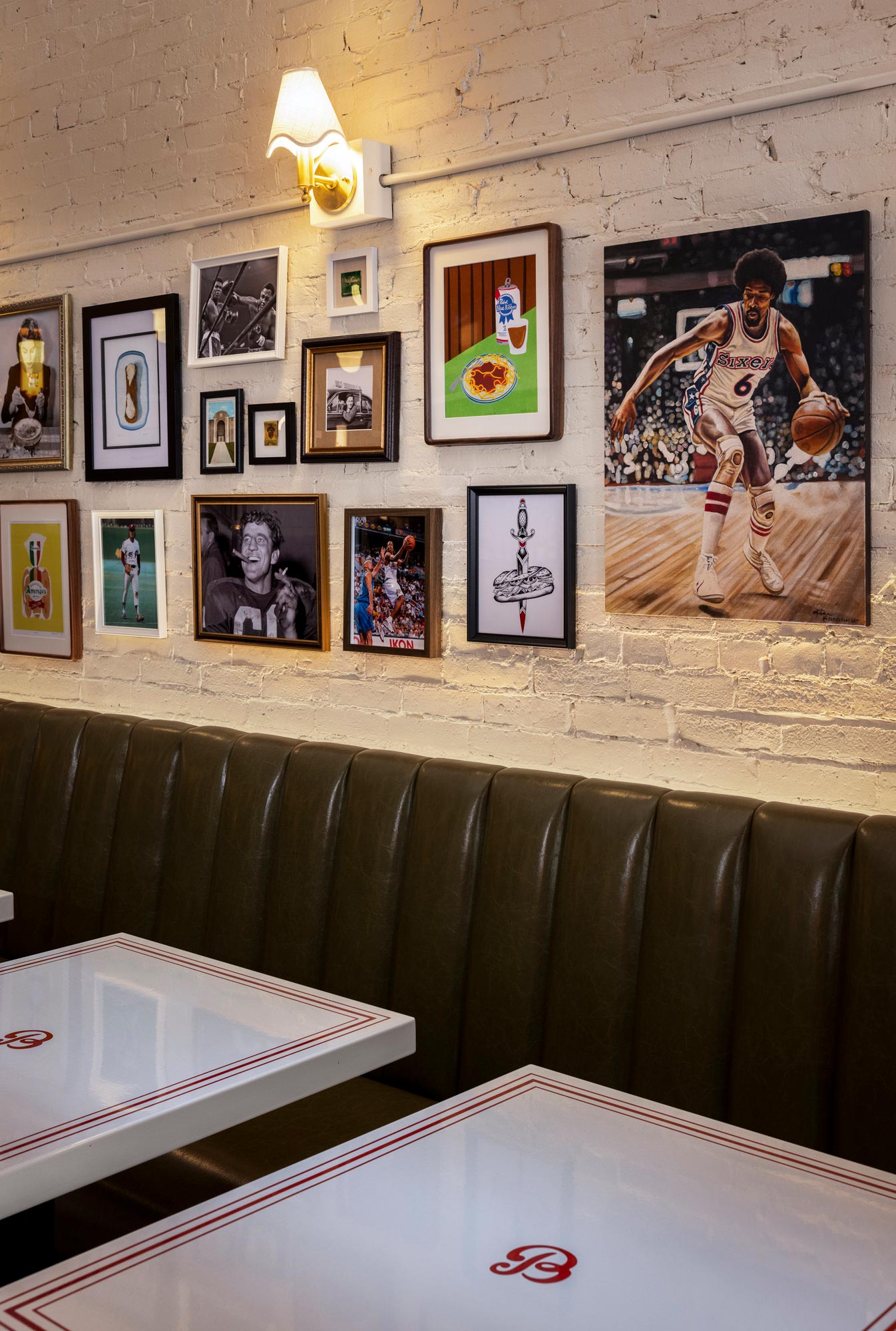 Framed Philadelphia and family-themed artwork displayed on a painted brick wall above a green leather banquette with white enamel tables featuring red pinstripes and a custom “B” logo at Benny’s Italian-American restaurant in Portland, Maine.