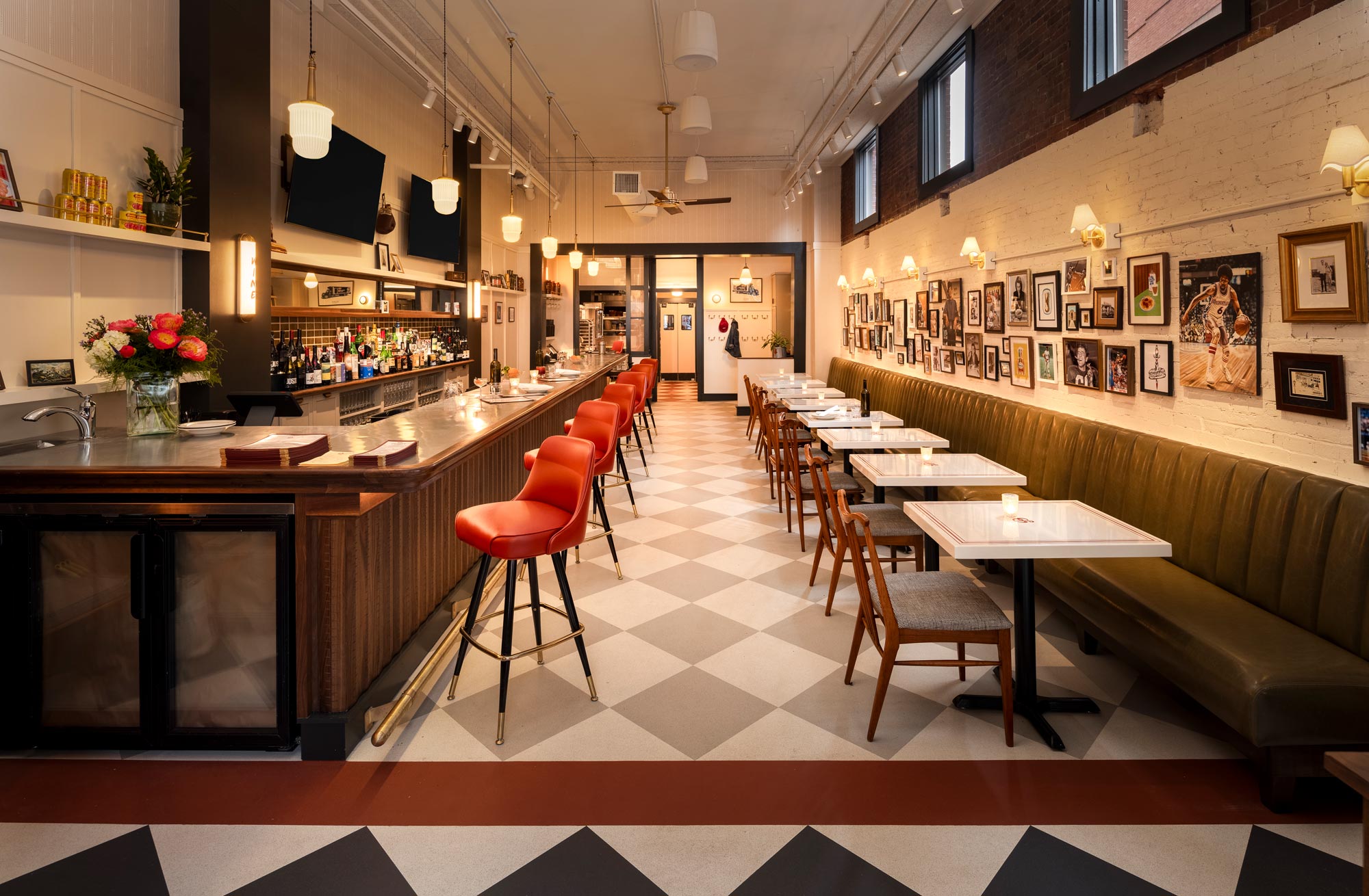 Full interior view of Benny’s in Portland, Maine, showing the checkered tile flooring, custom walnut and metal bar with red stools, green leather banquette seating, white enamel tables, gallery wall artwork, pendant lighting, and an open sightline to