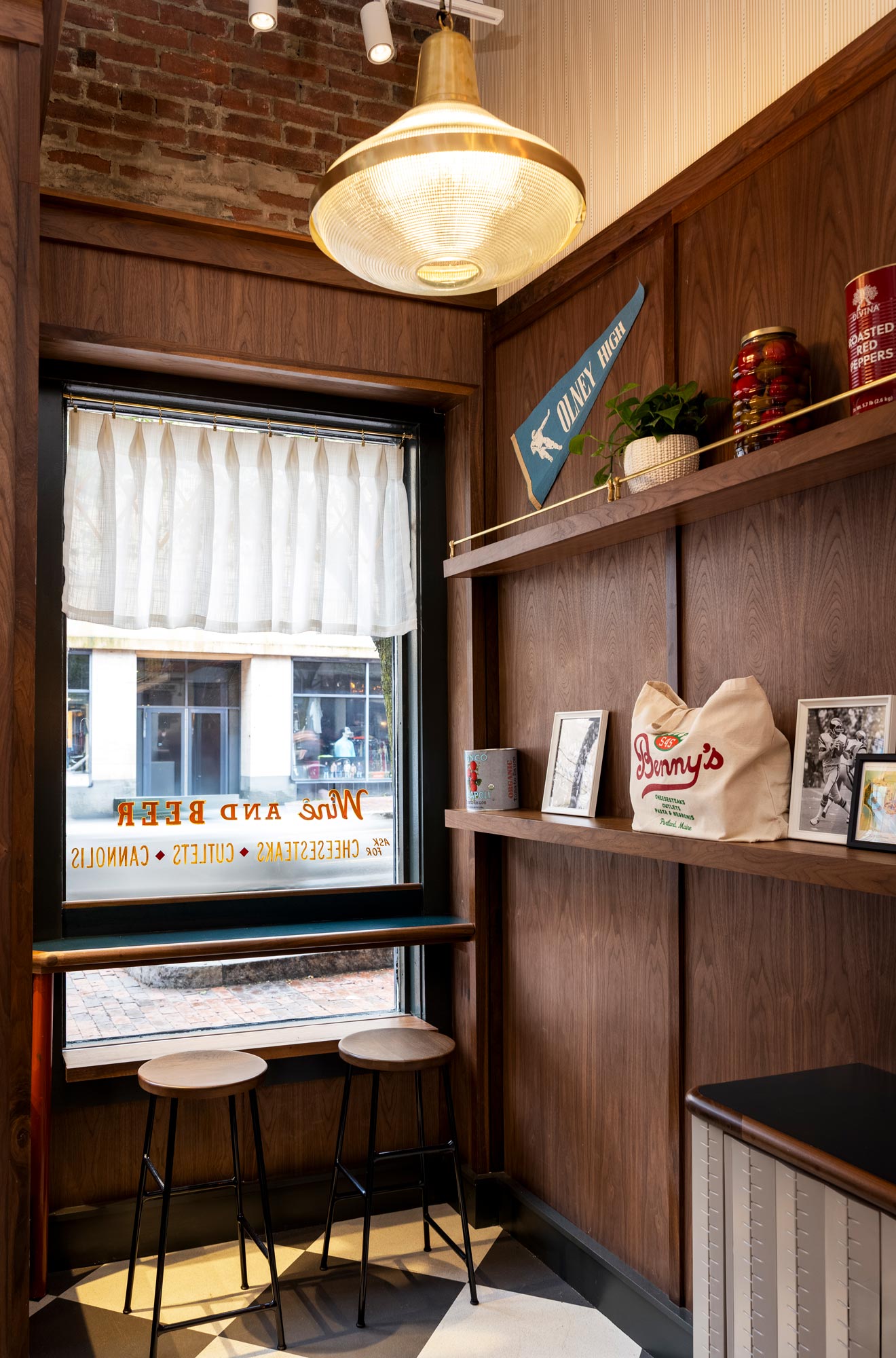 Window nook at Benny’s in Portland, Maine, with two wooden stools facing a street-view window framed by sheer curtains and vintage signage, adjacent to built-in walnut shelving displaying framed photos, branded merchandise, and preserved goods under