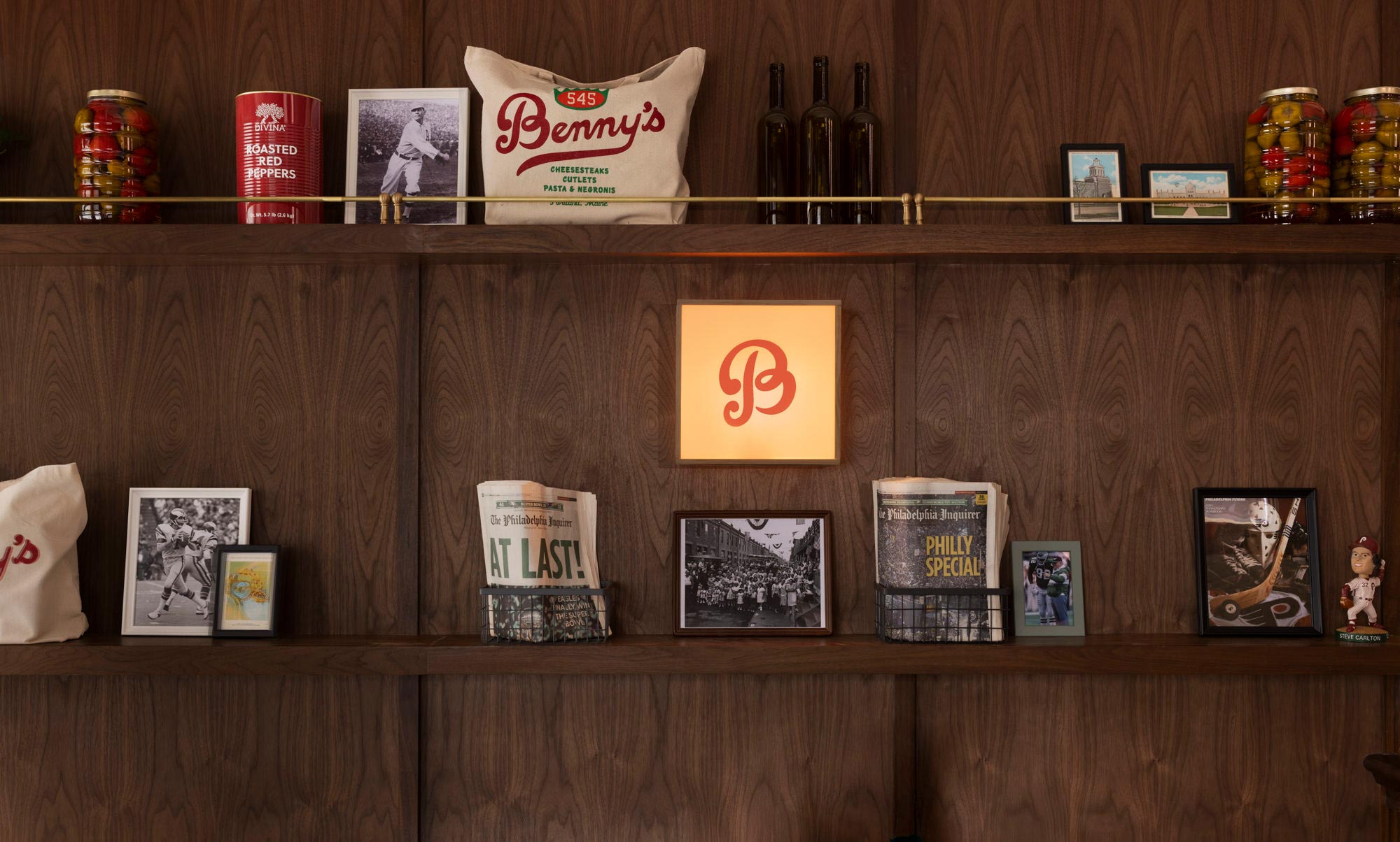 Close-up of the merchandise display at Benny’s in Portland, Maine, featuring framed vintage sports photos, Philly newspaper headlines, preserved peppers, a branded tote bag, and a glowing “B” logo sign, all arranged on custom walnut shelves with bras