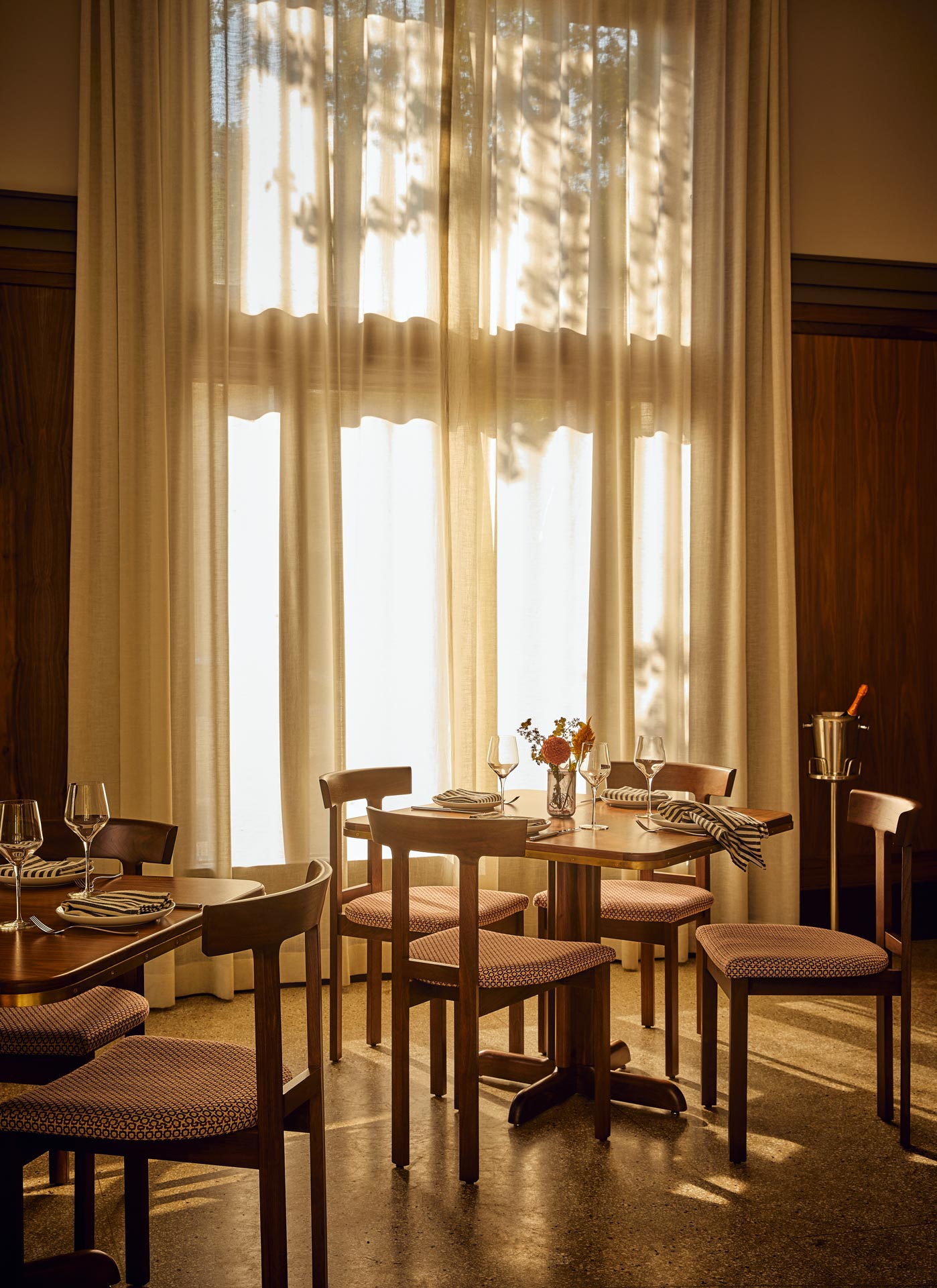 Sunlit dining area at Flat Iron Hotel in Asheville, North Carolina, featuring mid-century style wooden chairs, terrazzo floors, and sheer curtains casting soft shadows across the space.