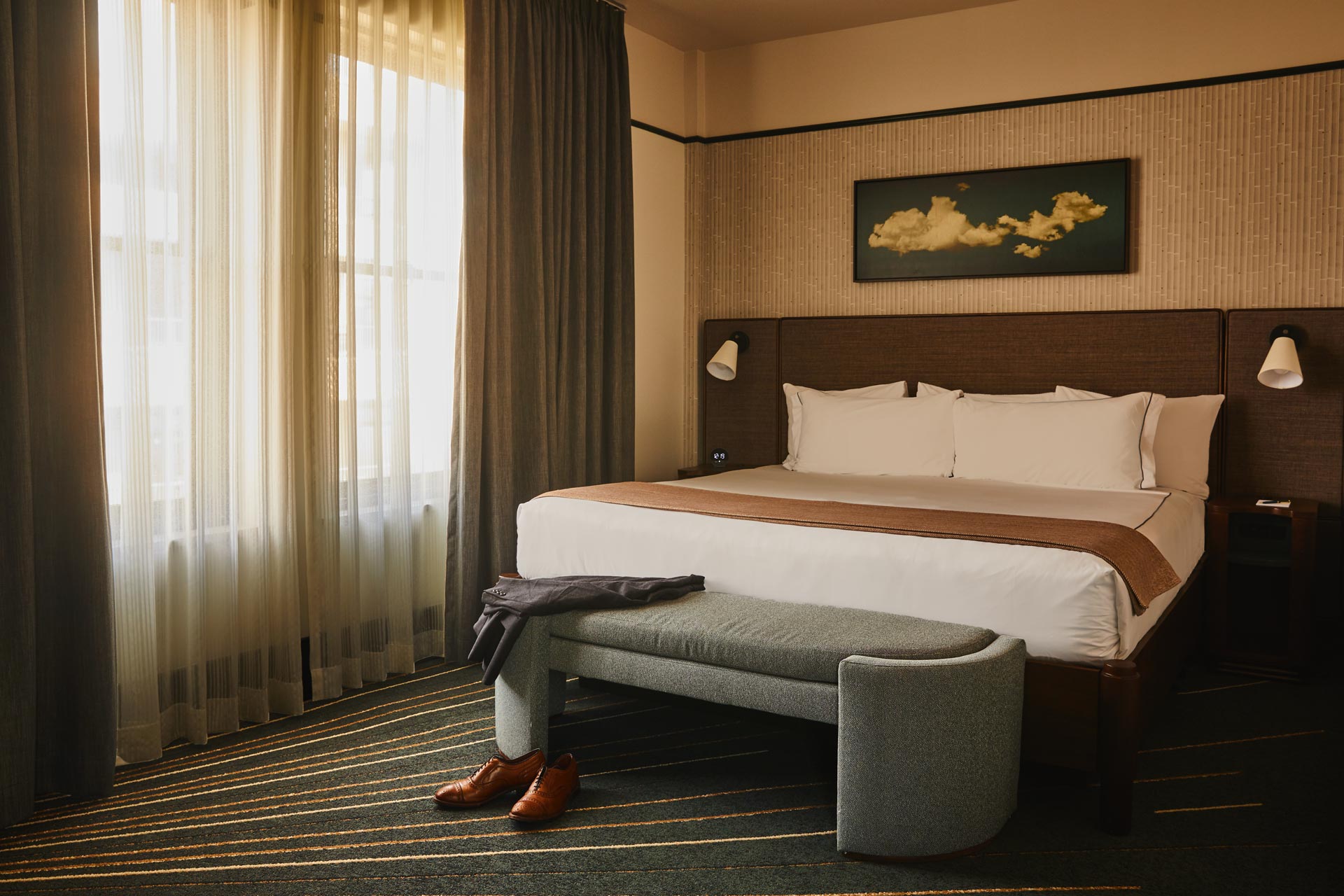 Flat Iron Hotel guest room with a king bed, upholstered bench, and framed cloud artwork, blending vintage-inspired textures and soft natural light through layered curtains.