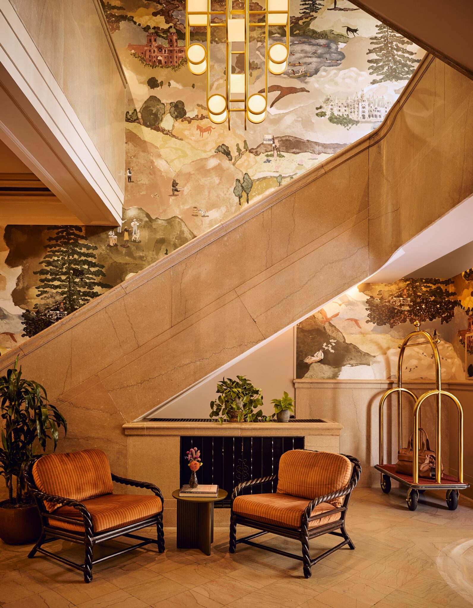 Lobby at the Flat Iron Hotel featuring a sweeping stone staircase, hand-painted mural, custom lighting, striped lounge chairs, and a brass luggage cart beside indoor plants.