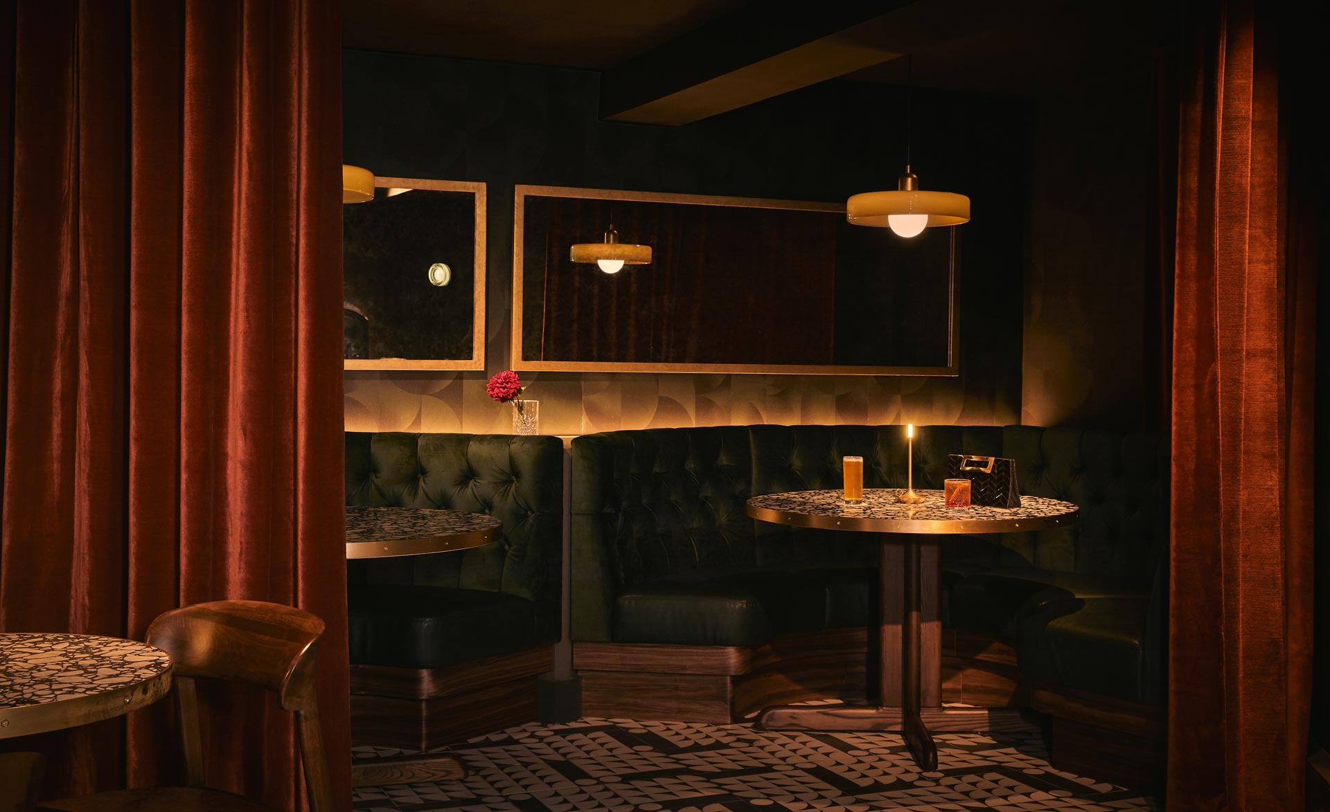 Intimate speakeasy seating at Flat Iron Hotel with tufted green velvet booths, patterned stone tables, soft globe pendant lights, and rich red velvet drapery.
