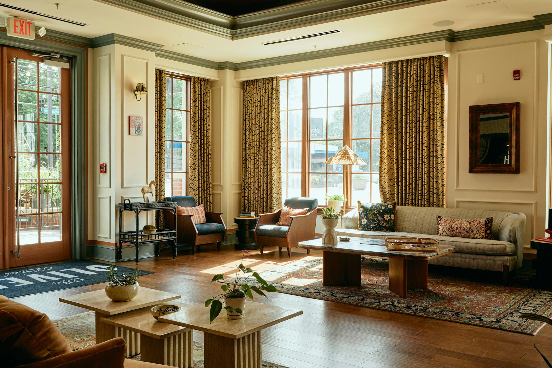 The Oliver Hotel of Oxford lobby with lounge seating, cream walls with green trim, and tall windows framed by mustard drapery with black geometric pattern. Warm wood floors with oriental rugs anchor seating areas of vintage-inspired furniture, including a sofa, armchairs, and low coffee tables decorated with small indoor plants. Natural light fills the boutique hotel’s interior, blending Southern tradition with modern design by Mey & Co.
